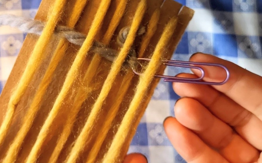 crafter weaves by passing paper clip shuttle through warp thread
