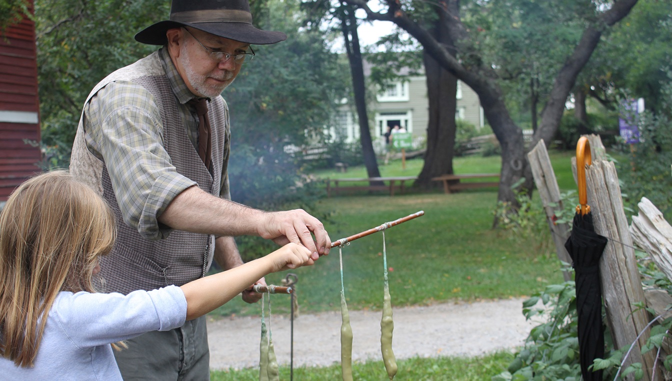 a costumed volunteer at Black Creek Pioneer Village shows a young visitor the traditional method of candle dipping
