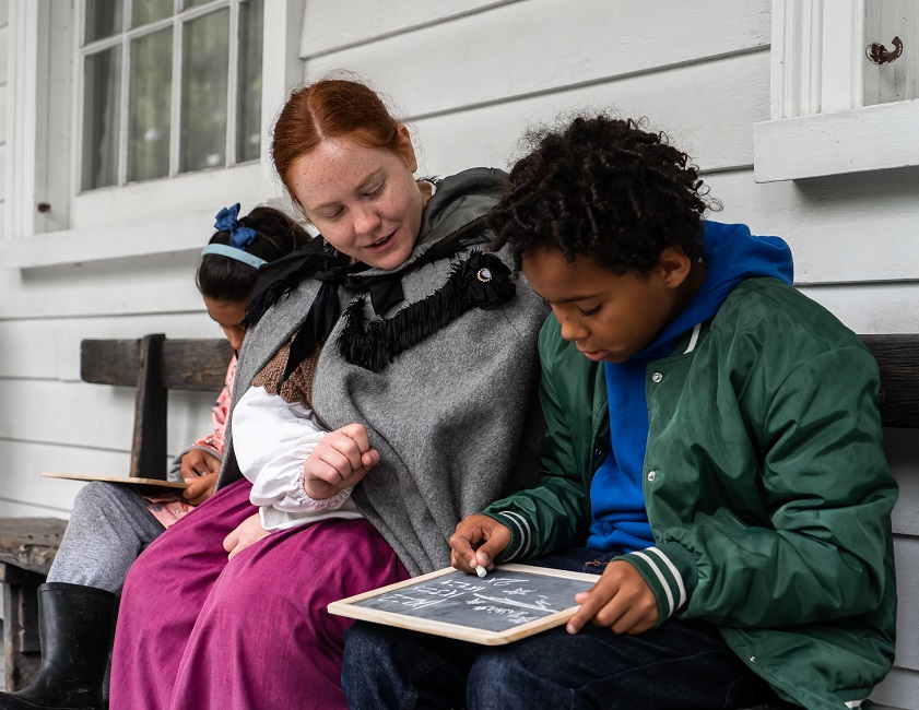 a costumed educator at the Village at Black Creek teaches young visitors how to use a 19th century slate to write out math problems