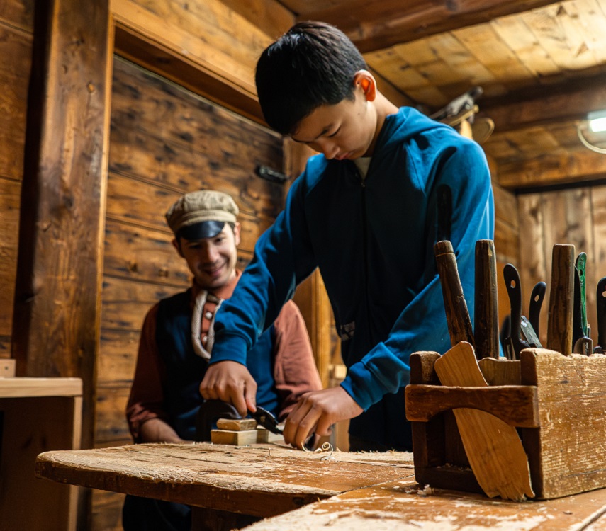 a student learns woodworking skill under the guidance of a costumed educator at the Village at Black Creek