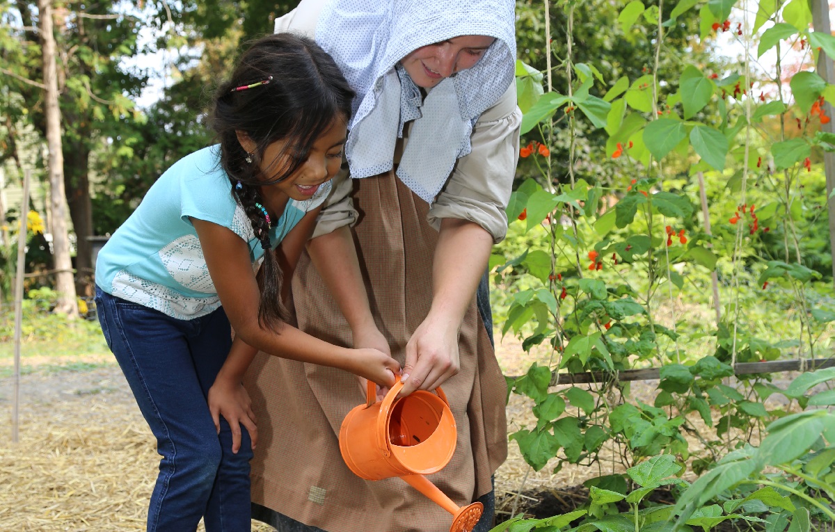 Summer Camp & Teen Experiences at The Village 13 a summer camper helps a costumed educator tend to a garden at The Village at Black Creek