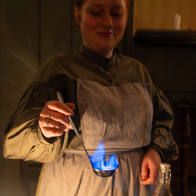 blue flame rises from a ladle as a costumed educator at The Village at Black Creek prepares a traditional Victorian festive concoction