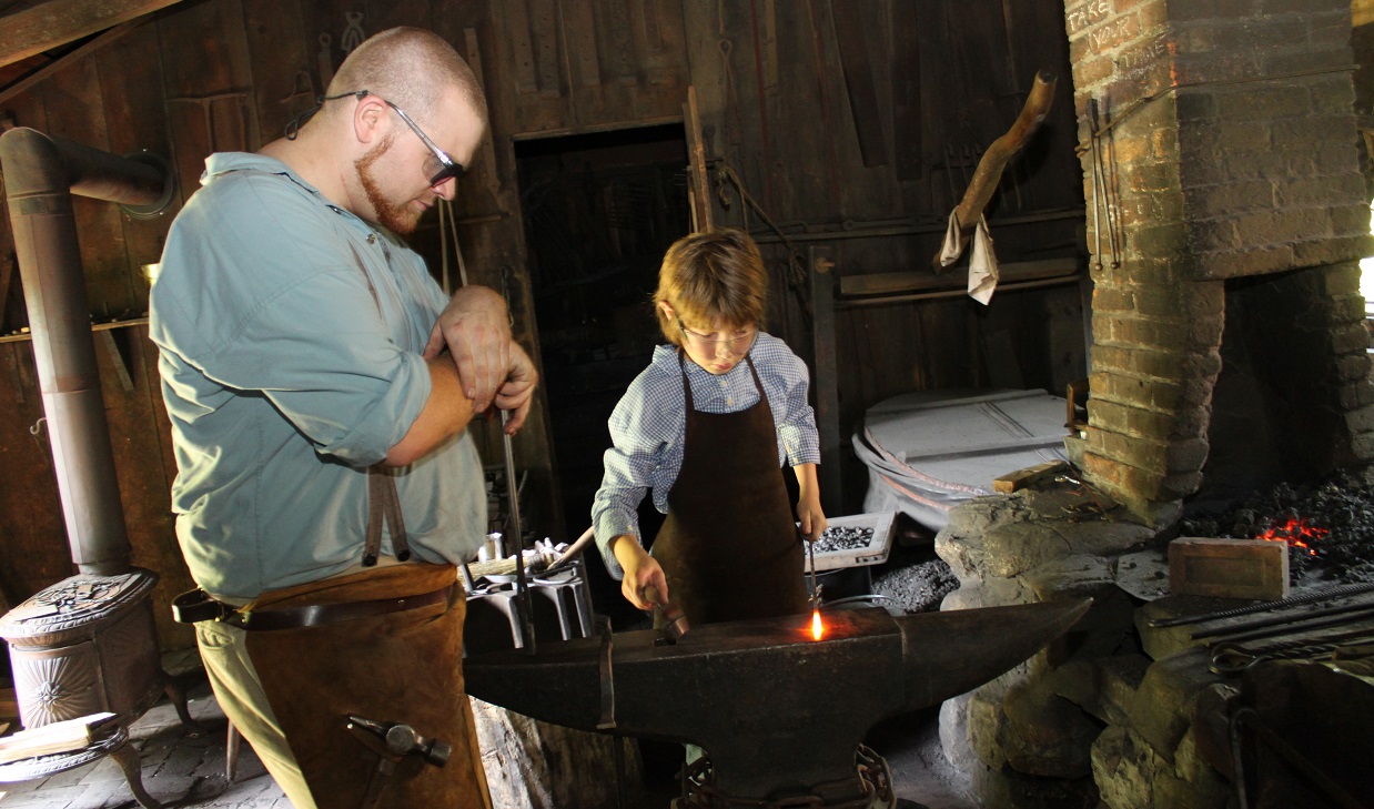 a blacksmith and his apprentice use an anvil to shape iron